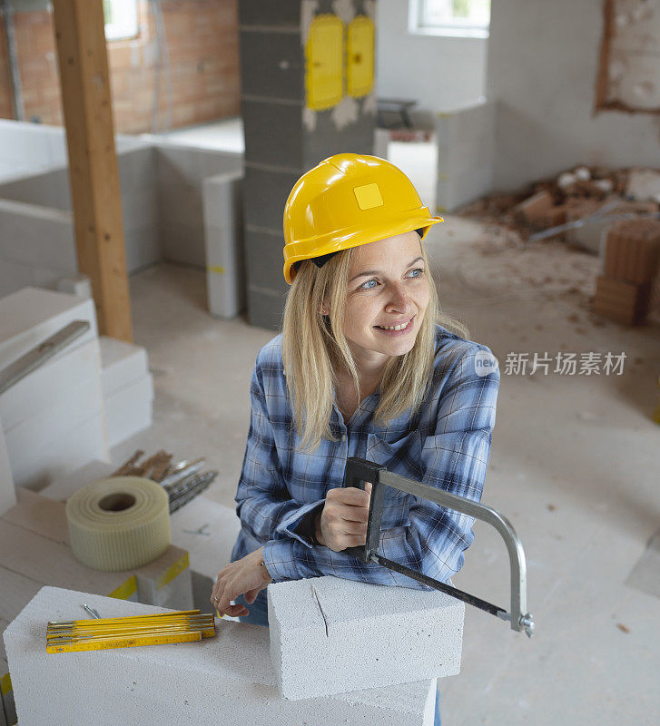 戴着黄色安全帽的年轻漂亮的女砌砖工正在建筑工地的房子里锯砖