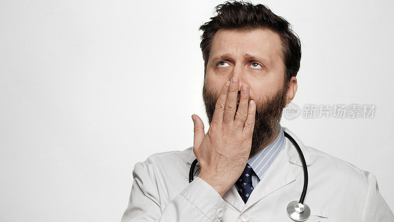 医生打了个哈欠。疲倦无聊的男人医生在白色的背景打呵欠，用手捂住他的嘴