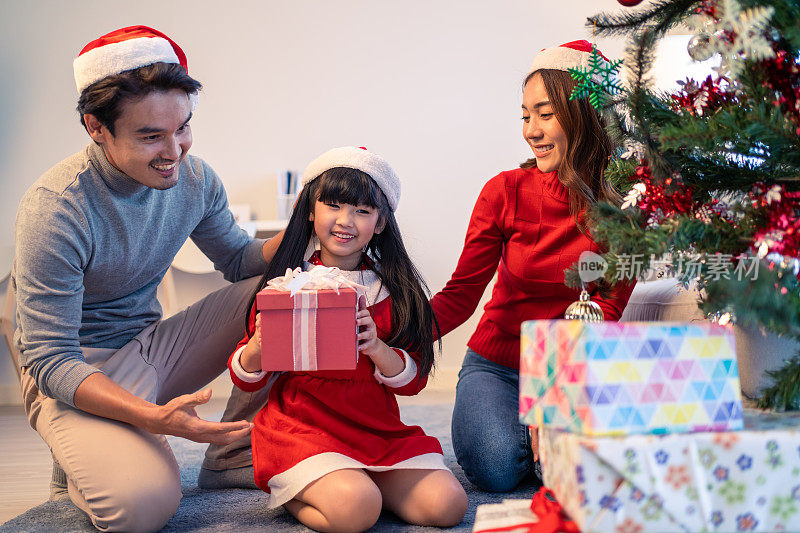 亚洲可爱的家庭成员用礼物装饰圣诞树。年幼的女儿心情愉快，兴奋地和父母一起在家里庆祝节日圣诞节感恩节聚会。