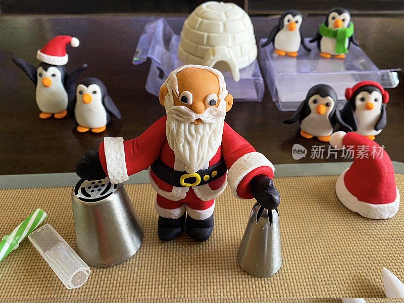 自制圣诞蛋糕顶饰(软糖糖霜)，圣诞老人，圣诞帽，白色的冰屋，戴着红色泡泡帽的企鹅，绿色的围巾和红色的耳套的特写图像