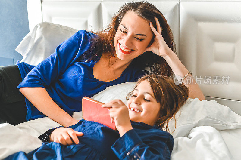 妈妈和女儿一起在床上放松地玩着，用手机看视频，拥有科技的幸福家庭