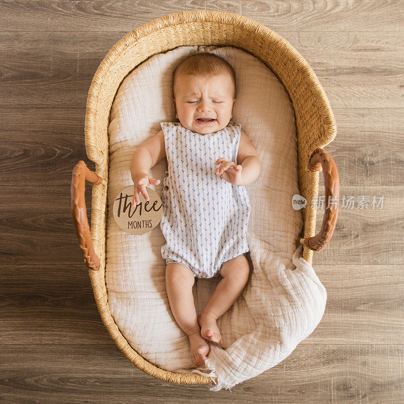 一个3个月大的男婴躺在一个舒适的奶油棉毯子里，躺在一个海草摩西篮子里哭泣