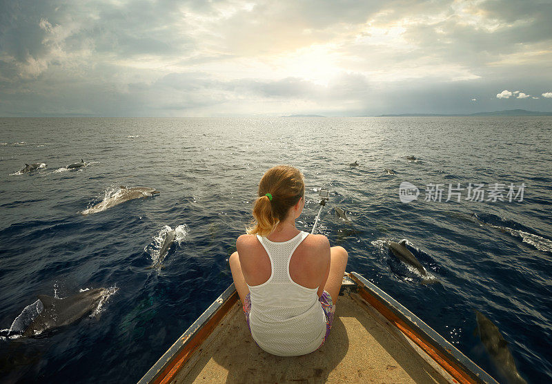 年轻的旅行者坐在船上拍摄一群海豚