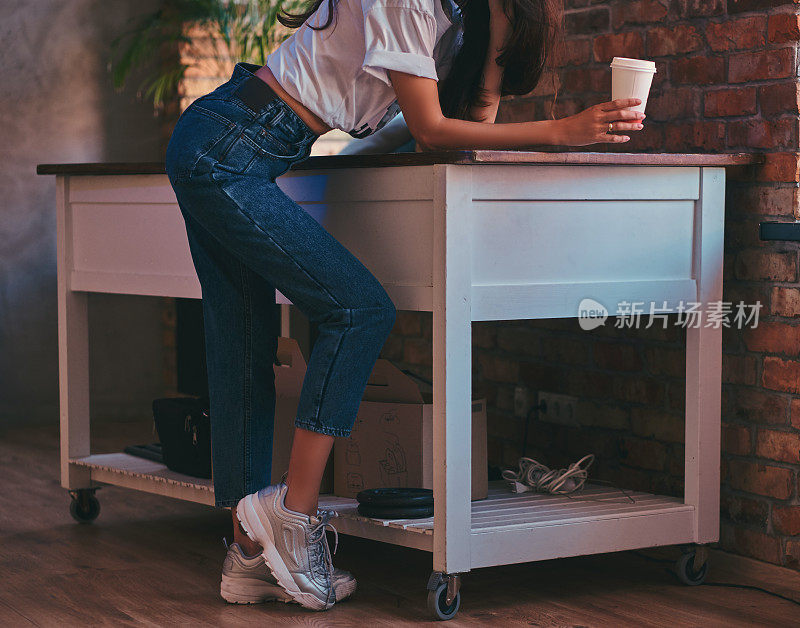 裁剪图像的一个很酷的现代女孩穿着白色的上衣和牛仔裤站在桌子上与一杯外卖咖啡在工作室阁楼内部。