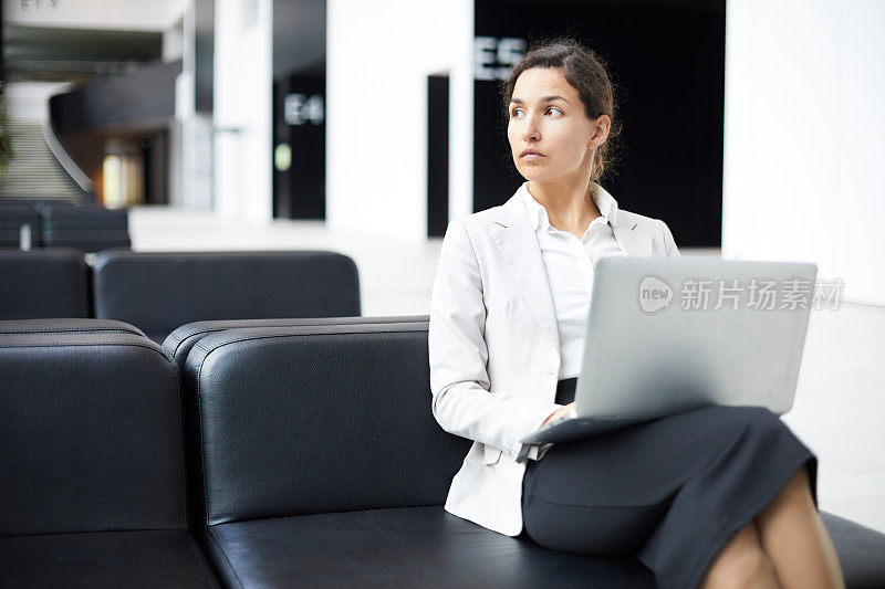 体贴自信的漂亮的混合种族女士在夹克坐在皮沙发和看别处，而使用笔记本电脑在大厅