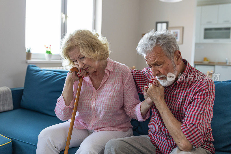 悲伤的退休妻子拄着拐杖坐在客厅里，旁边是老丈夫。老夫妇在家里手拉手看着对方