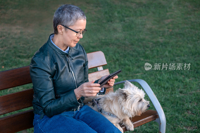一个成熟的女人和一只狗坐在长椅上，在平板电脑上阅读
