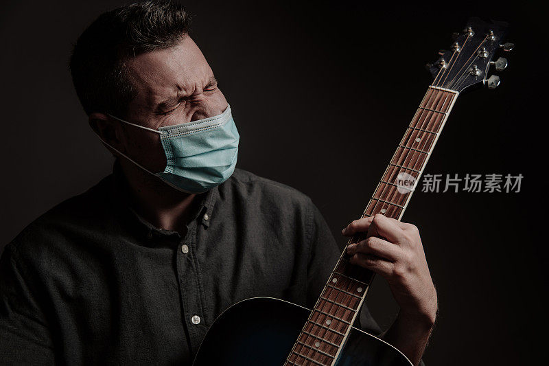 一个戴着医用口罩的男人站在黑暗的背景下，手里拿着一把旧吉他。