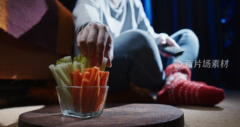 女人坐在地上，一边看电视一边吃芹菜和胡萝卜条