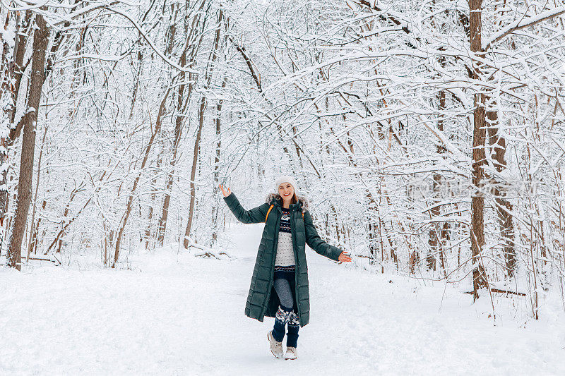 在冬天下雪的日子里，妇女在公园森林里散步，穿着绿色的蓝色皮夹克和帽子。快乐的独自旅行者探索当地的远足路线。