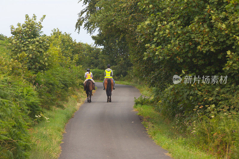 一对年轻的夫妇骑着马在威尔士乡村的小路上兜风，他们穿着亮黄色的高视镜安全装置，这样其他道路使用者就能看到他们。