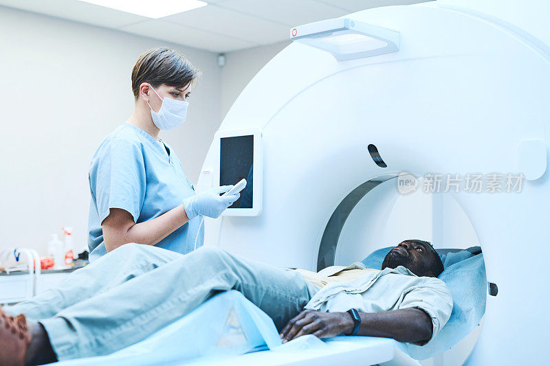 年轻的白人MRI技术人员在面具下按下控制面板上的按钮，同时为患者准备扫描