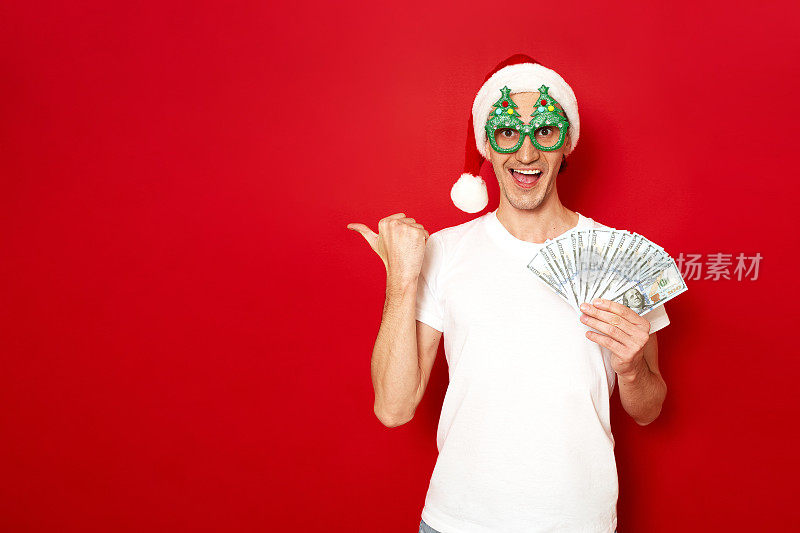 兴奋、高兴、惊讶的男子戴着圣诞老人的新年帽，手里拿着一叠美元钞票，手指着红色背景空间上孤立的自由空地上的文字概念——信贷、现金返还、银行、投资