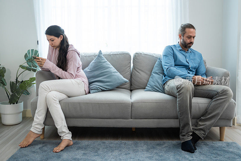 沉迷于手机的白人情侣不会注意对方。家庭问题，新婚男女坐在沙发上，用着智能手机，忽略家里客厅里的夫妻。