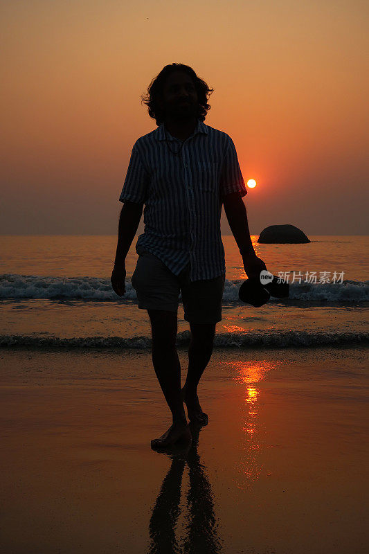 全帧图像的无法识别的人在果阿海滩度假，在日落时划水，Palolem，果阿，印度，度假游客赤脚走在海滩上，手里拿着鞋子在水里飞溅，橙色的日落，看着相机，聚焦在前景