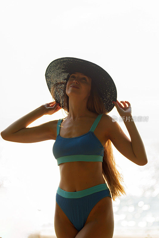 美丽的白人妇女在蓝色比基尼黑色沙滩帽和太阳镜在海滩上强烈的太阳背光穿过她的头