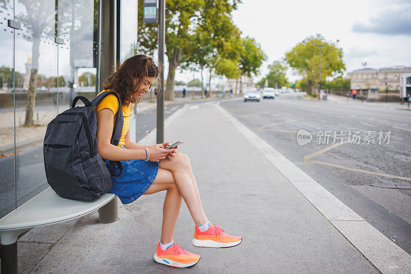 漂亮的年轻女子背着双肩包坐在公交车站玩手机