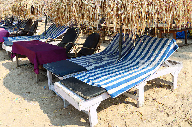 印度果阿，帕洛莱姆海滩，沙滩度假的特写照片，沙滩上有一排成对的蓝色软垫太阳躺床，茅草遮阳伞，蓝色和白色条纹的毛巾，边桌