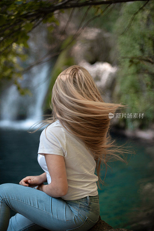 年轻女子坐在瀑布旁的大自然中，她的头发飘动着
