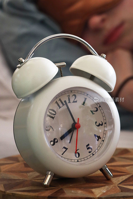 白色金属外壳复古闹钟的特写图像，双钟，白色表盘的模拟显示时钟坐在桌子旁边的木头上，红头发的女人戴着睡眠面罩在双人床上，躺在枕头上睡觉，重点是前景