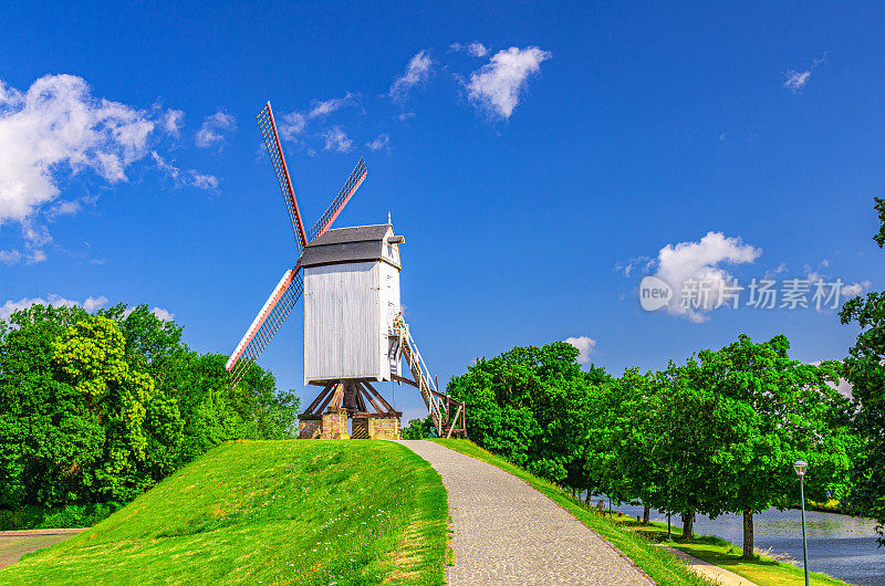 比利时佛兰德地区，布鲁日市靠近河流的绿色山丘上的传统老风车，夏日的蓝天，典型的带有风帆的白色老式木制风车和绿树成荫的公园小径