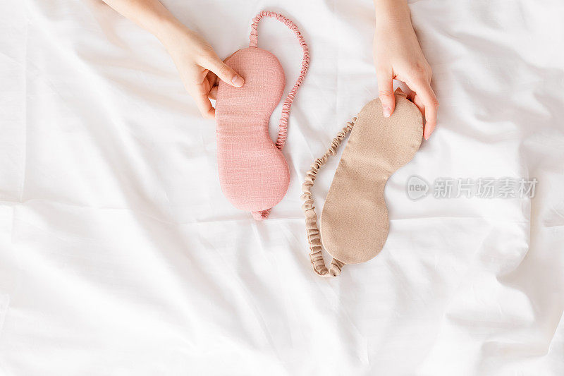 合上两个睡眠用的眼罩，米色和粉色放在白色的被褥上，女人手拿一个，尽量平躺。俯视图睡眠面膜，最佳睡眠者，舒适放松。好好休息的概念。
