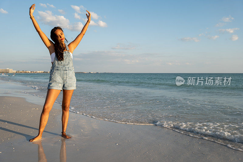 一个快乐的女人在沙滩上，在日落时分，在海边散步，张开双臂