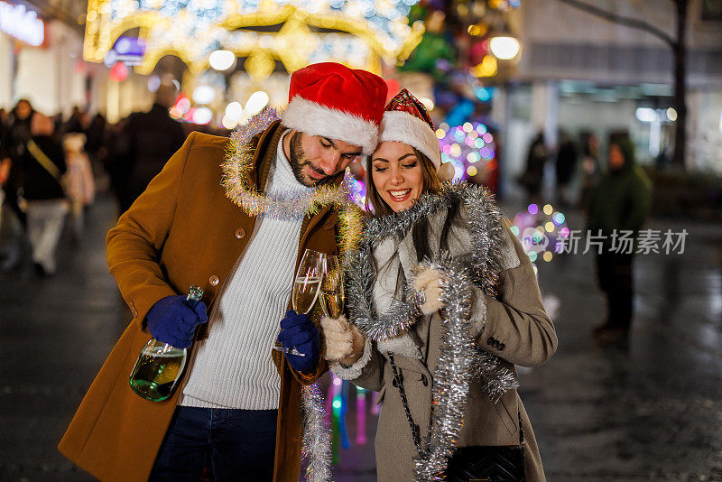 一对戴着圣诞帽的年轻夫妇在城市的街道上饮酒作乐。