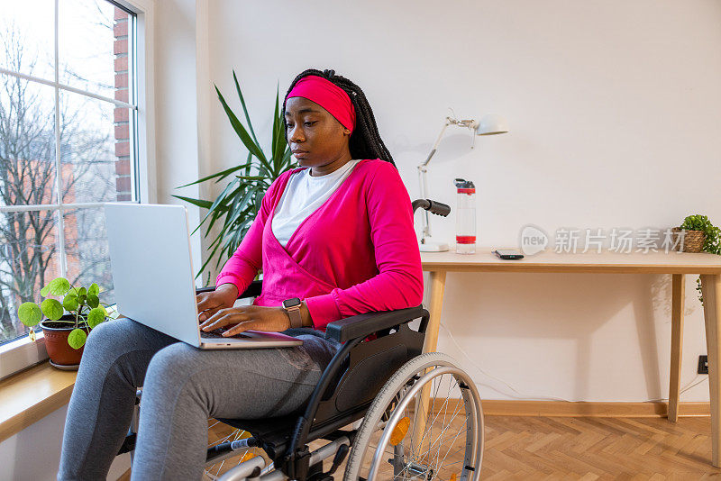 黑人妇女坐在轮椅上用笔记本电脑工作，靠窗