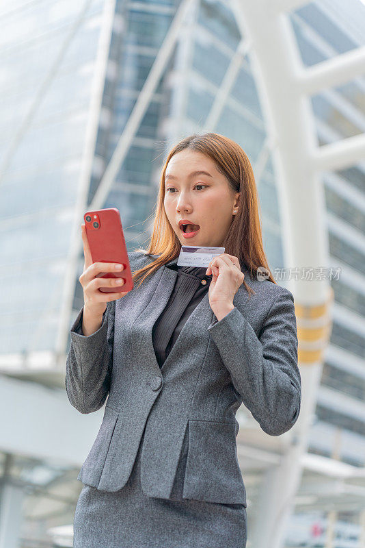 一名年轻的亚洲女商人穿着灰色的办公套装，在商业区用她的智能手机使用信用卡进行在线交易，她微笑着自信