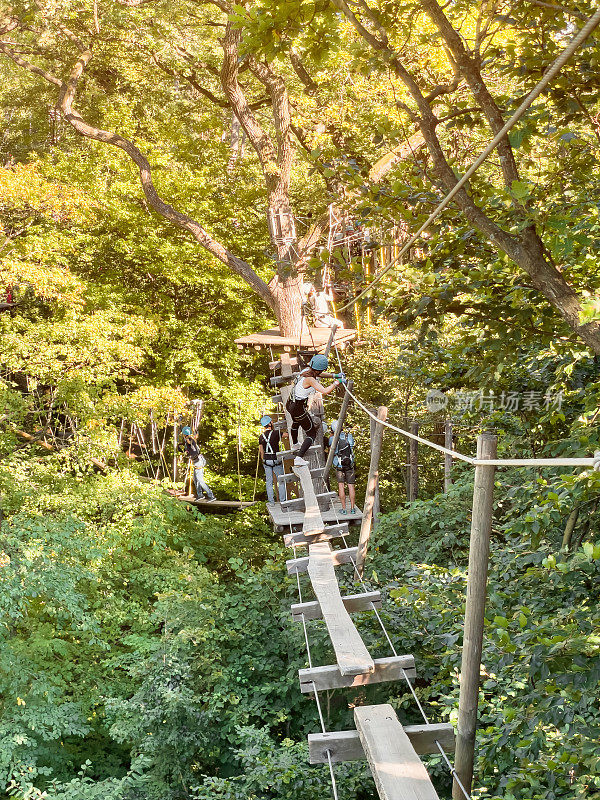 夏日里，一个十几岁的女孩穿着安全装备和头盔，在一个攀岩冒险公园里沿着树间的索桥散步。木林中的绳索公园。冒险公园特写。夏季的乐趣和运动冒险的人。