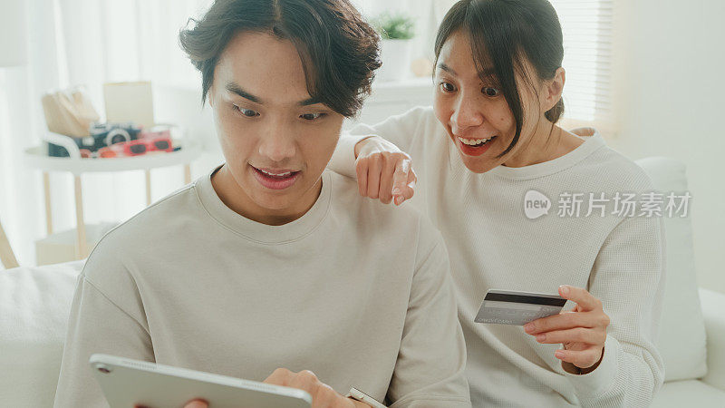 年轻的亚洲夫妇坐在家里客厅的沙发上，一起玩电子平板电脑购物，使用信用卡在线支付。生活方式花时间在家里。