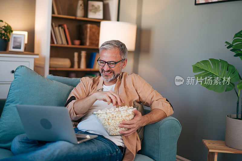 快乐的家伙坐在家里的沙发上，用笔记本电脑看体育比赛，吃着爆米花。