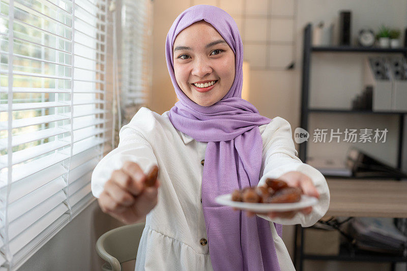 快乐的年轻亚洲穆斯林妇女戴着紫色的头巾，一只手拿着一盘干枣子，另一只手拿着一个枣子，对着镜头微笑