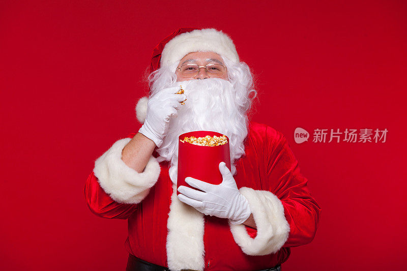 传统的圣诞老人看电视，吃爆米花。圣诞节。红色的背景。情感恐惧惊喜