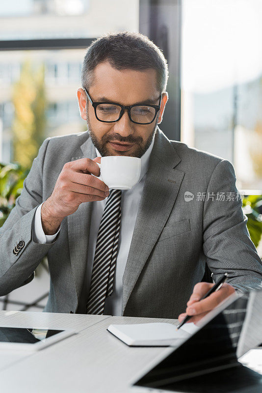 西装革履，戴眼镜的商人，喝着咖啡，在工作的地方写着笔记