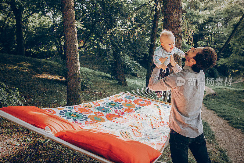 快乐的潮爸爸和他的儿子在公园附近的吊床上玩，而在森林露营，家庭的概念，英俊的男人抱着婴儿在户外