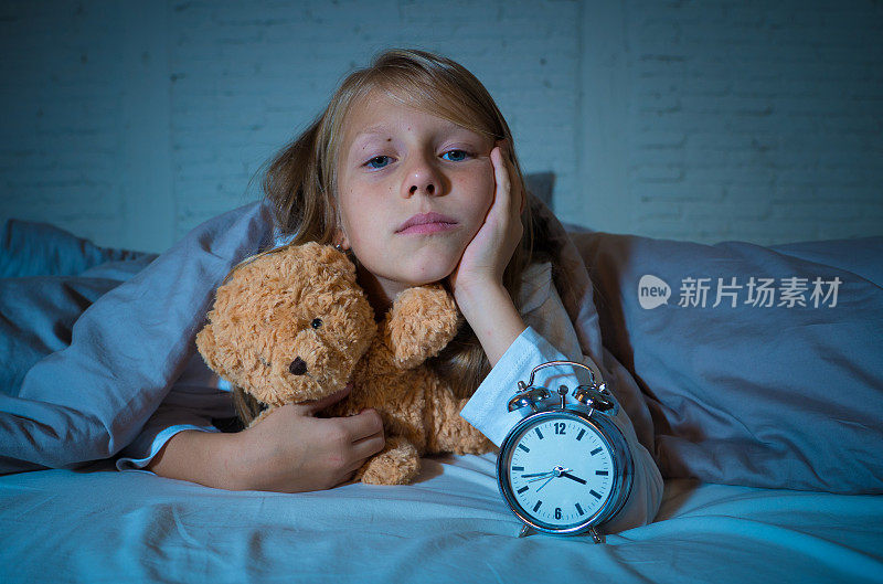 可爱的失眠小女孩躺在床上，闹钟看起来很累，有睡眠问题，晚上睡不着觉，或在儿童失眠、焦虑和睡眠障碍中过早醒来。