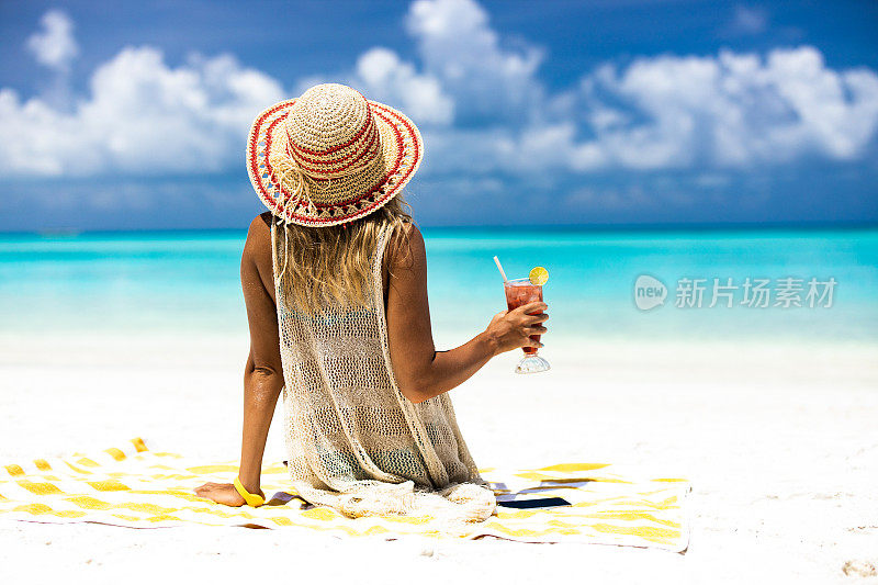 一个放松的女人与鸡尾酒在海滩上的后视图。