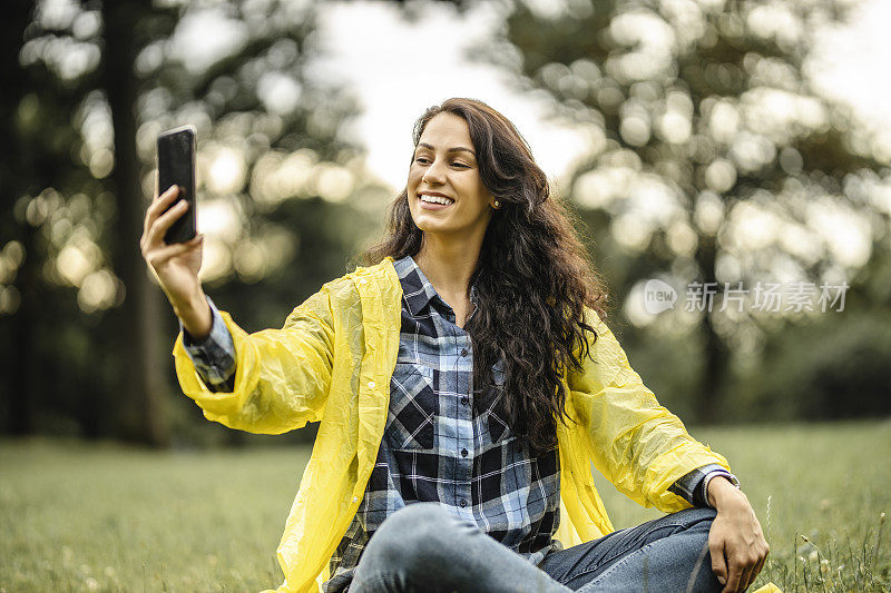 美丽的年轻女子在黄色雨衣与她的手机自拍在自然