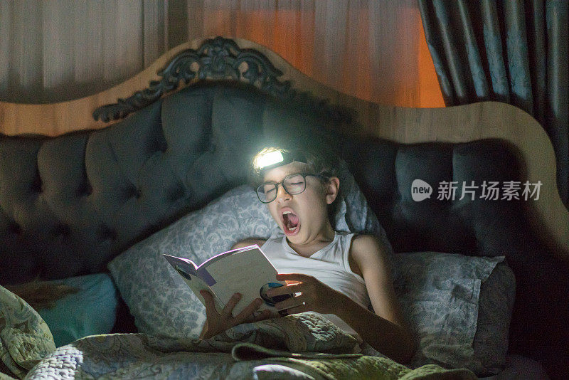 小男孩一边看书一边在床上打哈欠。