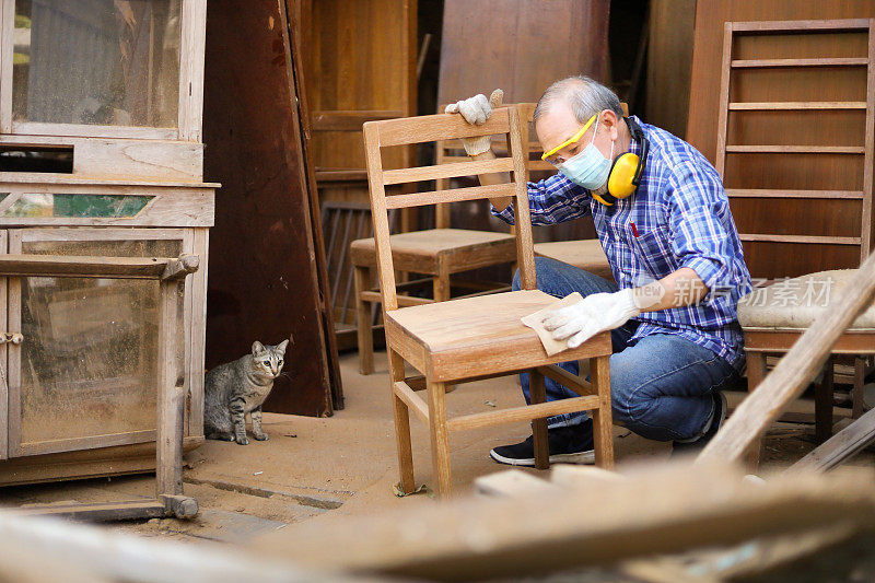 一位年长的亚洲木匠正在他自己的车库风格的工作室里打磨木椅，他的猫在一旁看着