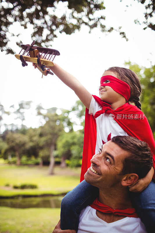 穿着超级英雄服装的女孩坐在父亲的肩膀上，手里拿着玩具飞机