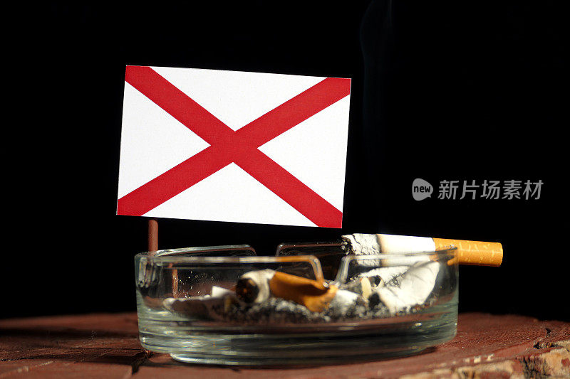 阿拉巴马州国旗和燃烧的香烟在烟灰缸孤立的黑色背景