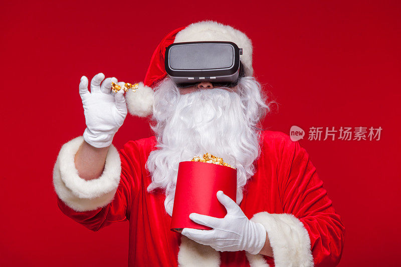 圣诞老人戴着虚拟现实眼镜和一个装满爆米花的红桶，在红色的背景上。圣诞节
