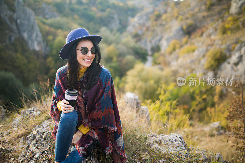 女旅行者一边喝着咖啡，一边欣赏着山景。一个年轻的女游客喝着一杯热饮，欣赏着山上的风景。徒步旅行的概念