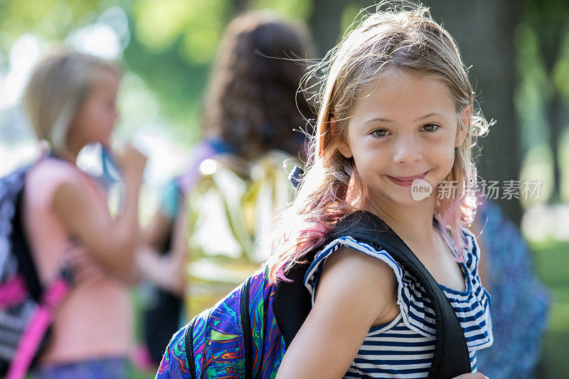 可爱的小学女孩在镜头前微笑，背着书包在校园里