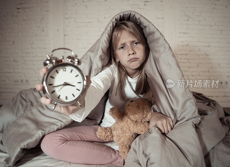 可爱的失眠小女孩坐在床上，显示闹钟看起来很累，有睡眠问题，晚上睡不着或醒得太早在儿童失眠，焦虑和睡眠障碍。