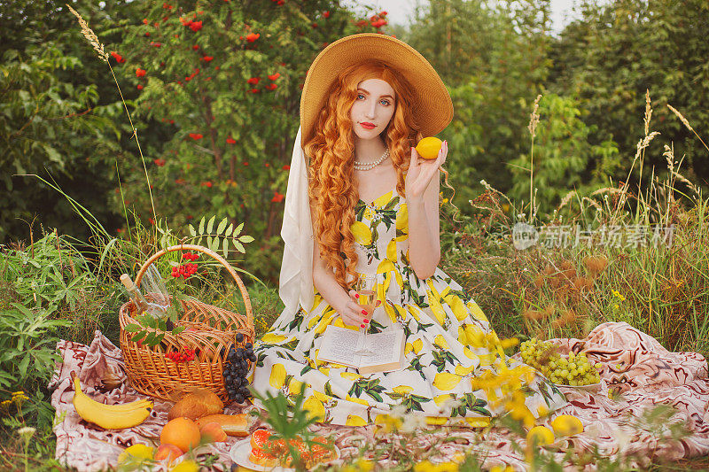 红发鬈发的女孩，穿着夏季连衣裙，床上放着野餐篮。戴着帽子的女人，背景是夏天的自然。漂亮的金发模特。夏季野餐。美味的食物