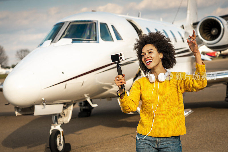 一名年轻的女视频博主在一架停在机场跑道上的私人飞机旁用自拍杆装着相机，正在做一场秀